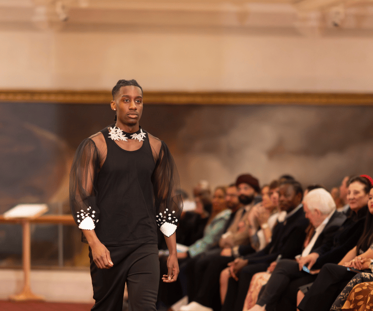 Male model walks the runway wearing a black outfit with sculptural hand accessories, at Fashion Frenzee by Central London Alliance, part of London Fashion Week, in front of a seated audience at Guildhall Art Gallery