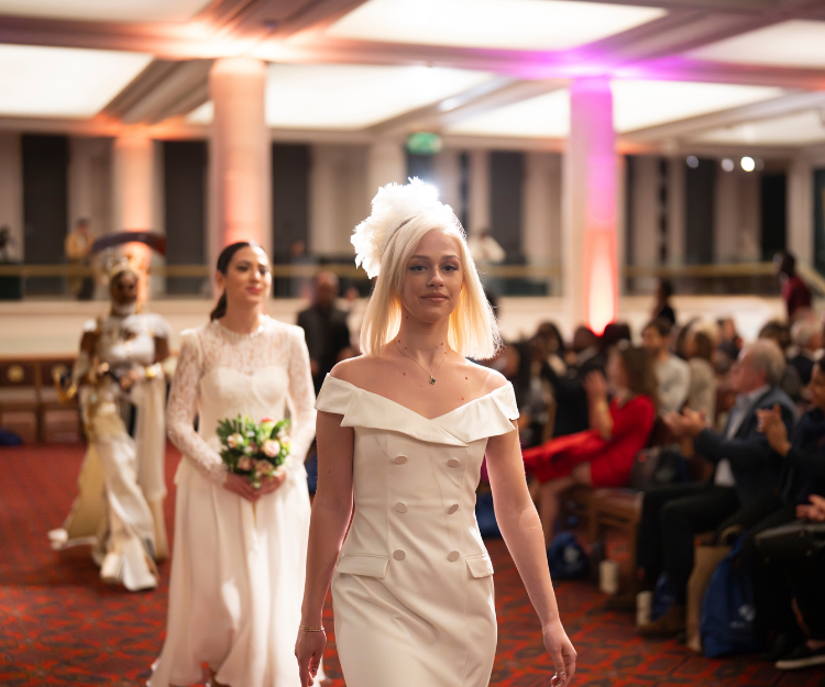 Two models walk the catwalk wearing white bridal-style gowns, one holding a bouquet, at Fashion Frenzee by Central London Alliance, part of London Fashion Week, showcasing pre-loved fashion
